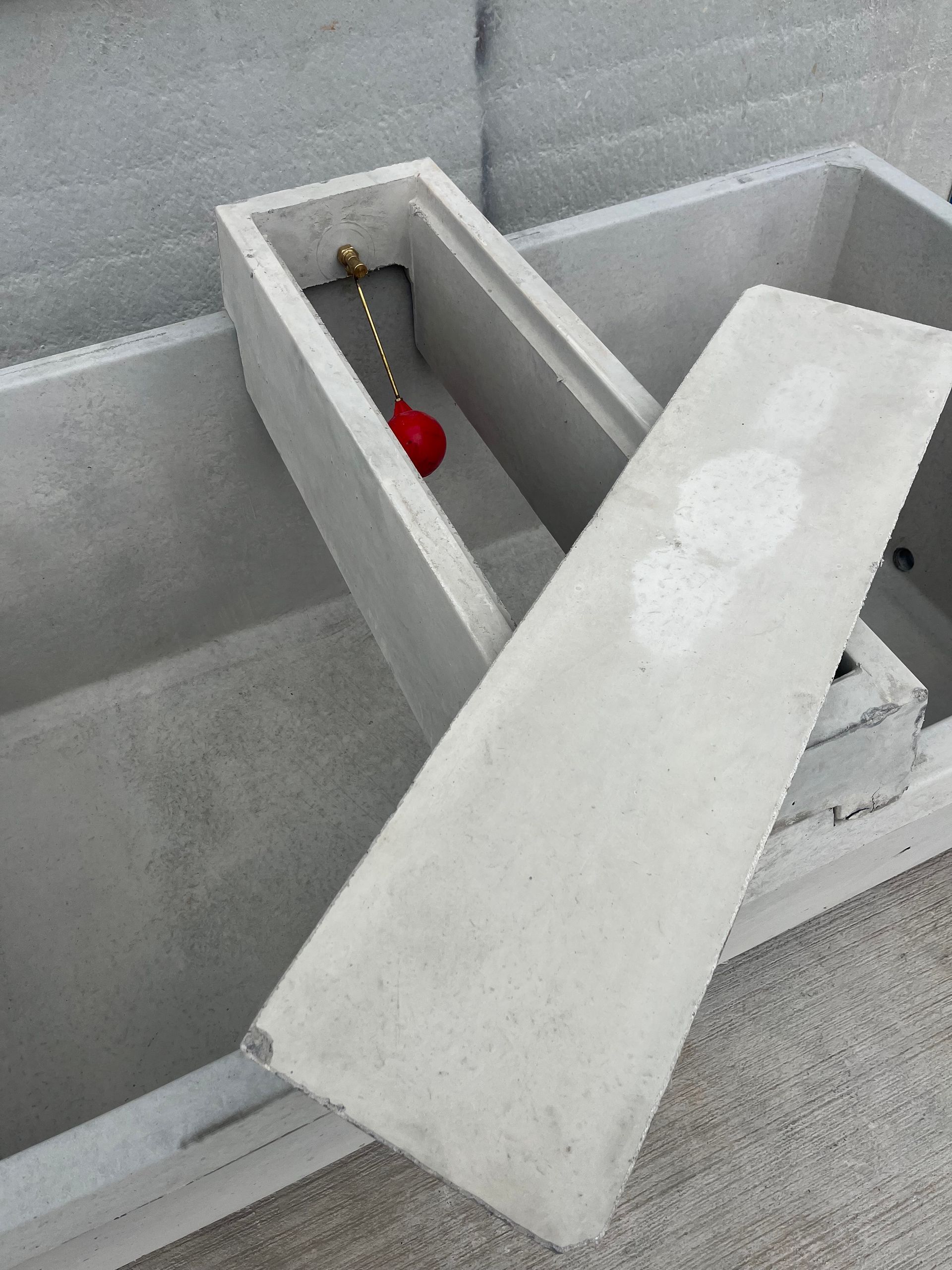 A concrete livestock trough with a detached lid and a red float valve assembly installed in the end compartment.