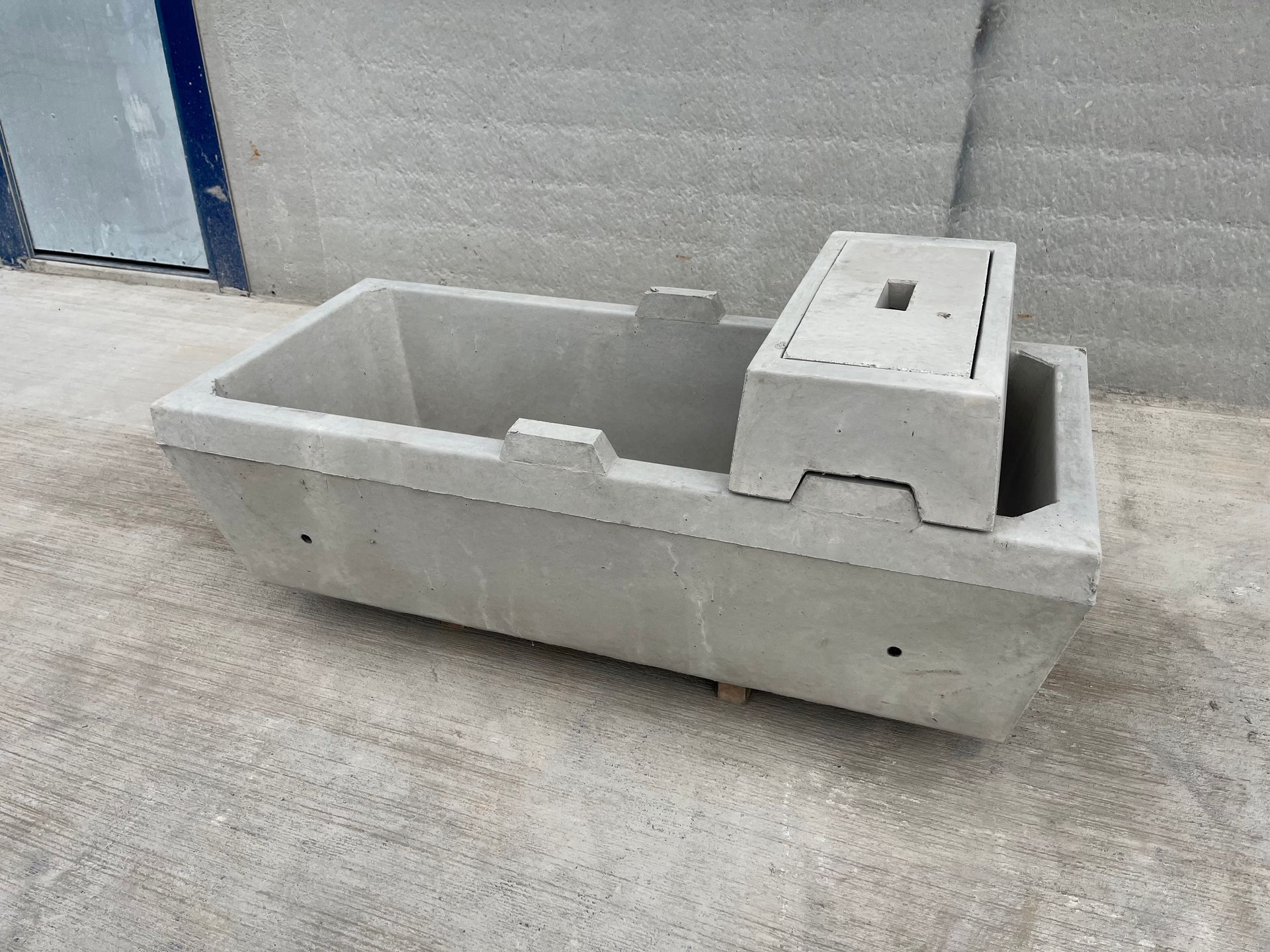 A grey concrete cattle water trough with a covered utility section, resting on a concrete floor.