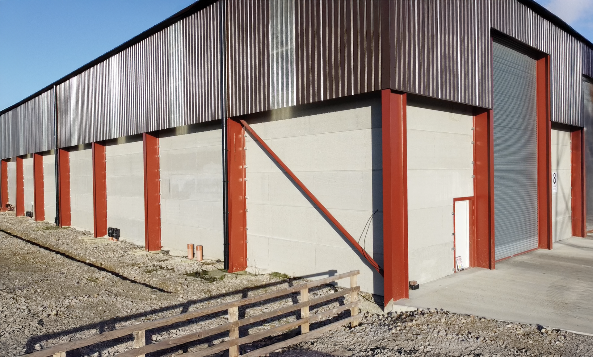 A warehouse exterior featuring red steel support beams, light grey concrete lower walls, and corrugated metal siding.