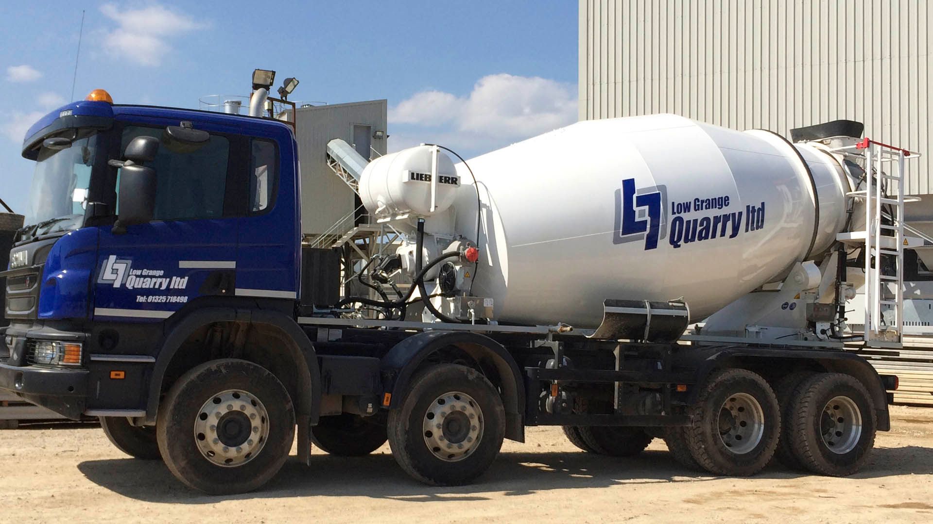 A blue and white industrial cement mixer truck parked on a gravel lot under a clear blue sky.