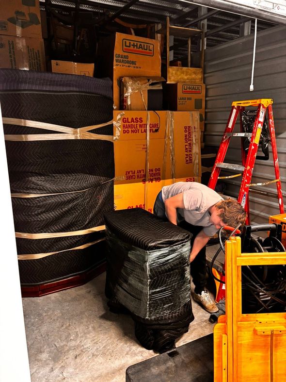 Warehouse worker bending over a pallet of stacked boxes beside a red ladder and yellow forklift