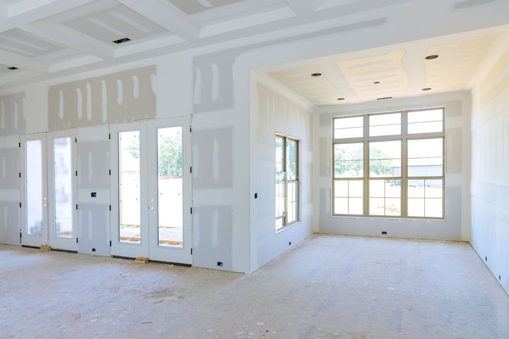 Person in yellow hard hat applying joint compound to drywall with two trowels.
