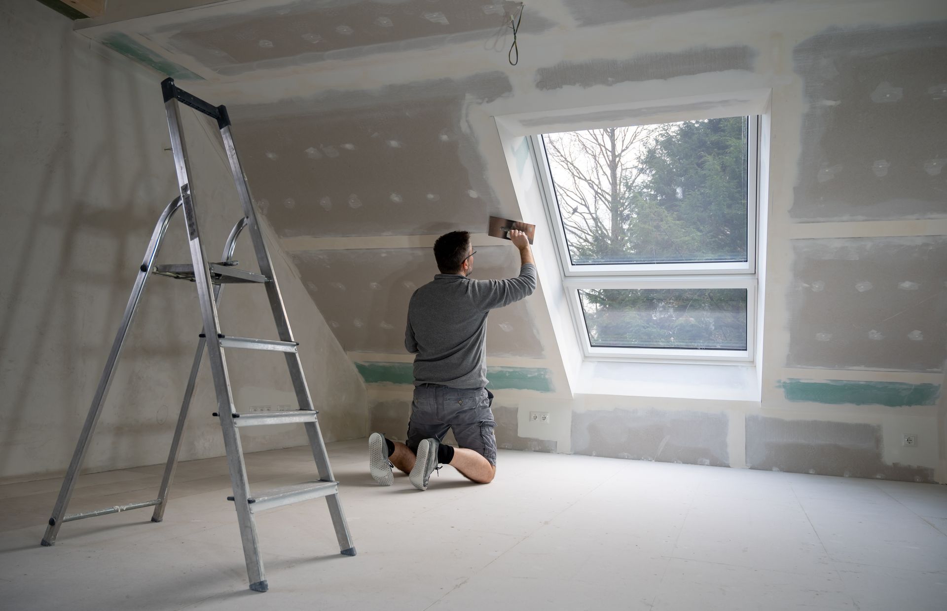 Construction worker on ladder, installing drywall in a framed interior space.