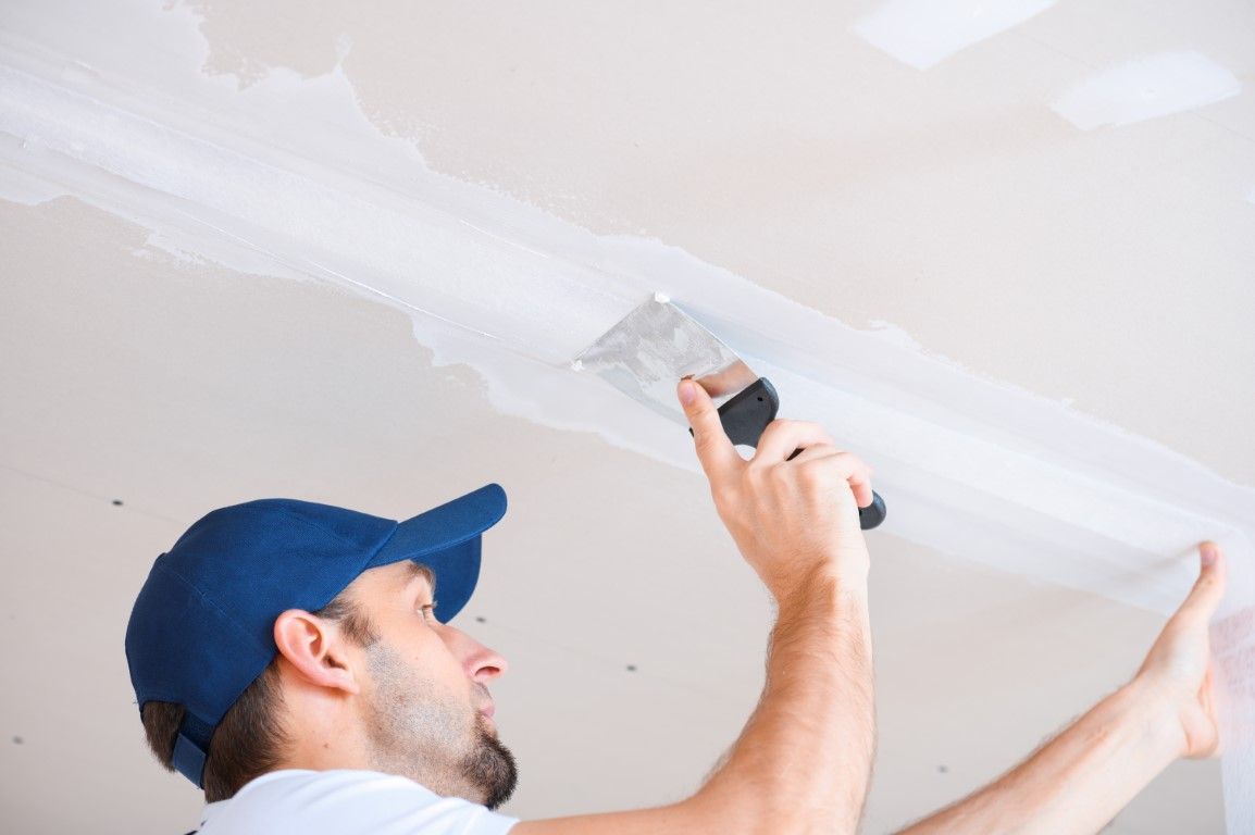 Man in blue cap applies joint compound to a ceiling with a trowel.