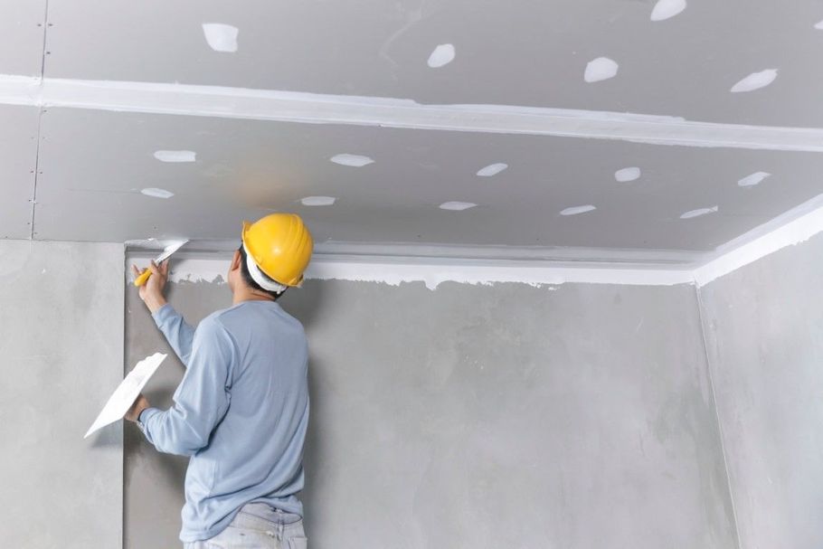 Person in hard hat applying drywall compound to ceiling joints with a trowel.