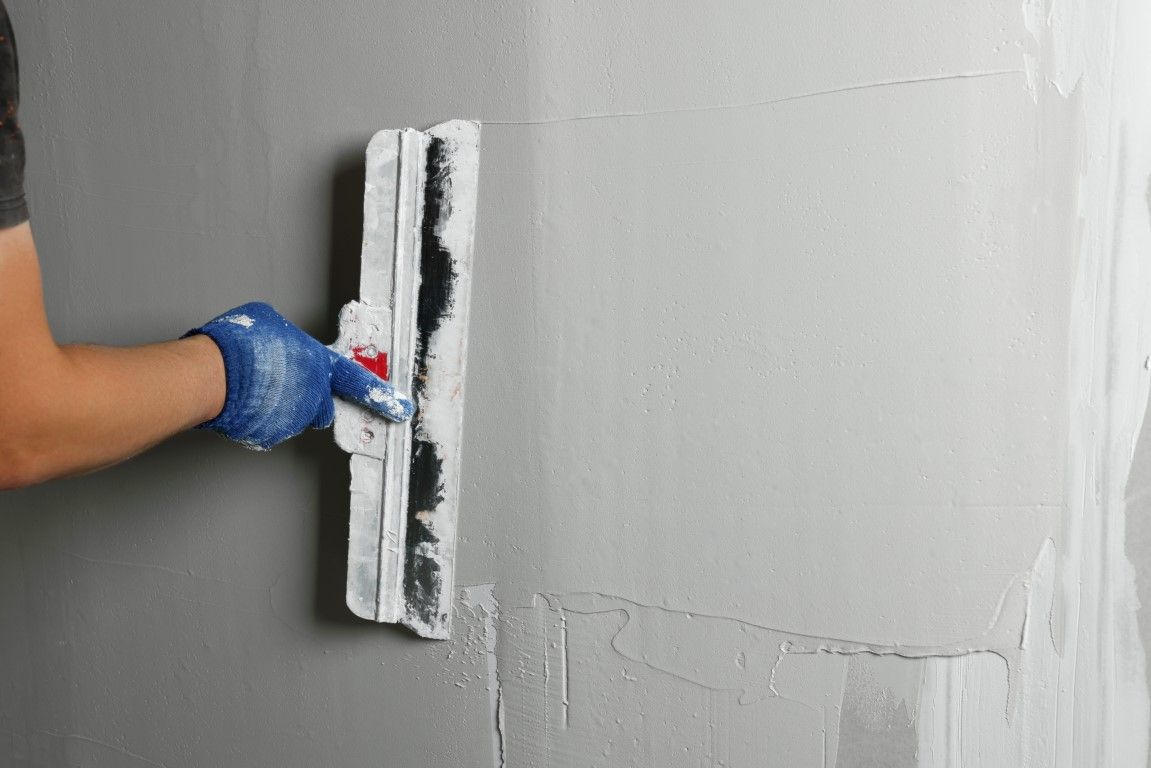Person in blue glove using a trowel to apply light-colored plaster on a gray wall.