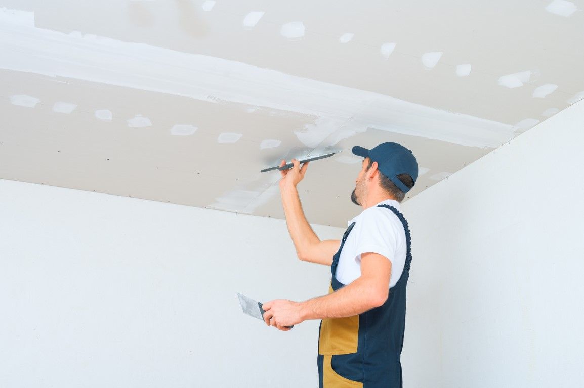 Man applying drywall compound to a ceiling with a trowel, wearing a blue cap and overalls.