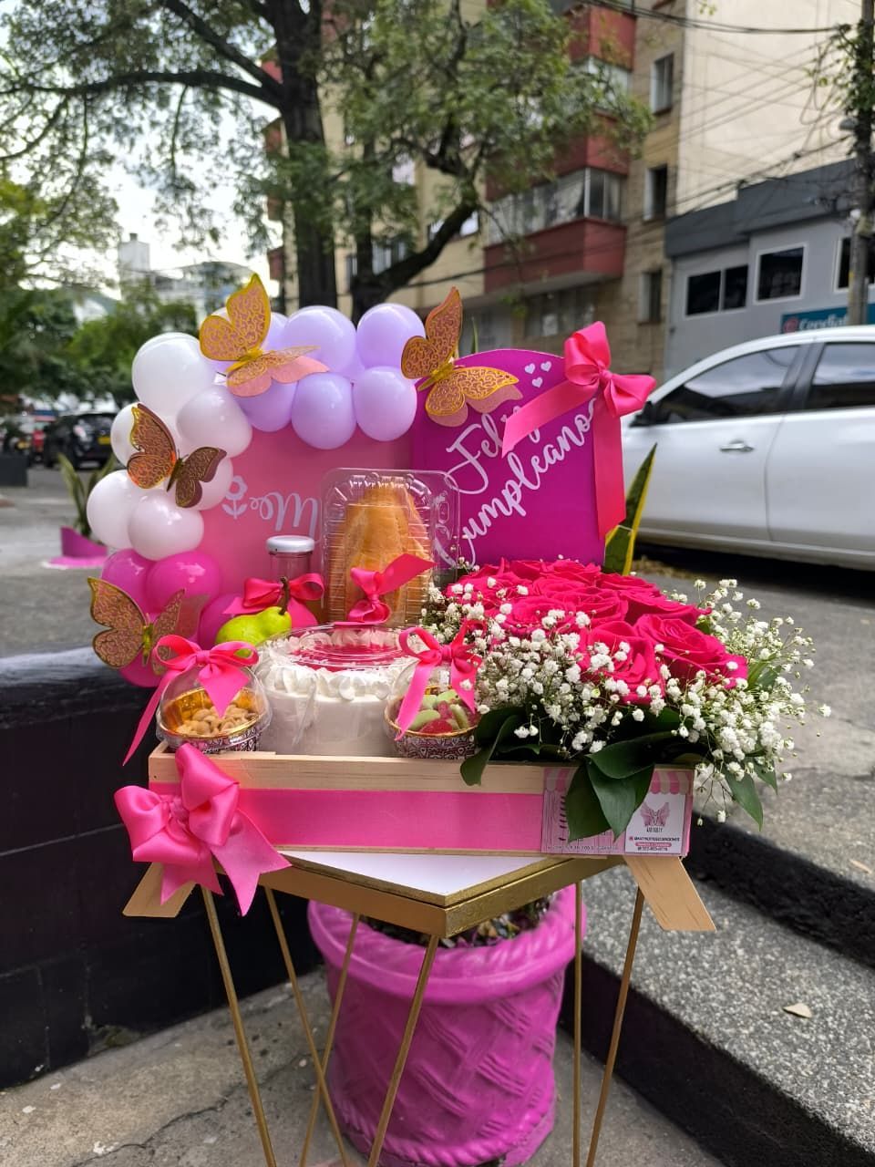 Cesta de regalo de cumpleaños rosa con flores, globos y dulces sobre un soporte.