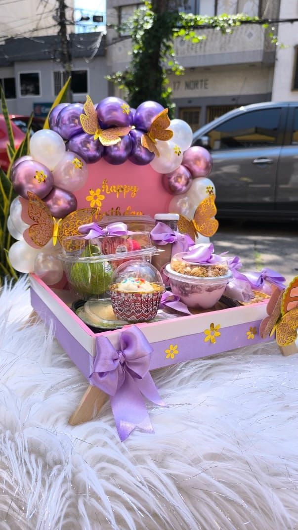 Bandeja de desayuno con decoración morada y dorada, que incluye globos, lazos y recipientes para comida.
