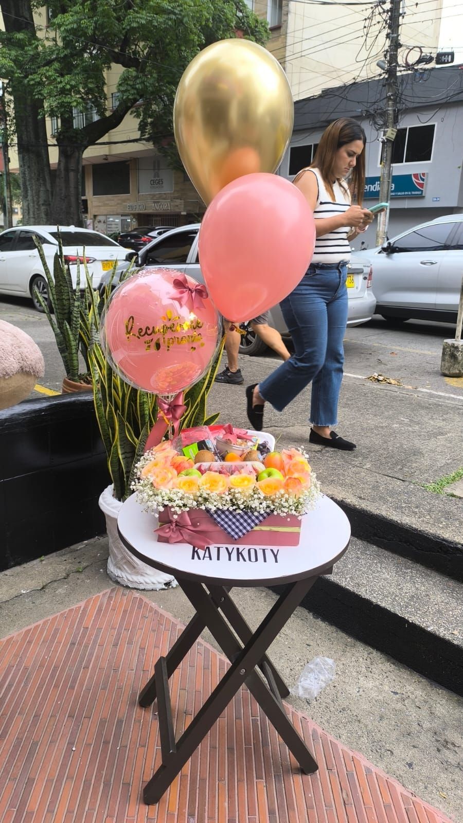 Mesa con un arreglo floral y globos, cerca de una persona usando su teléfono. Al aire libre.