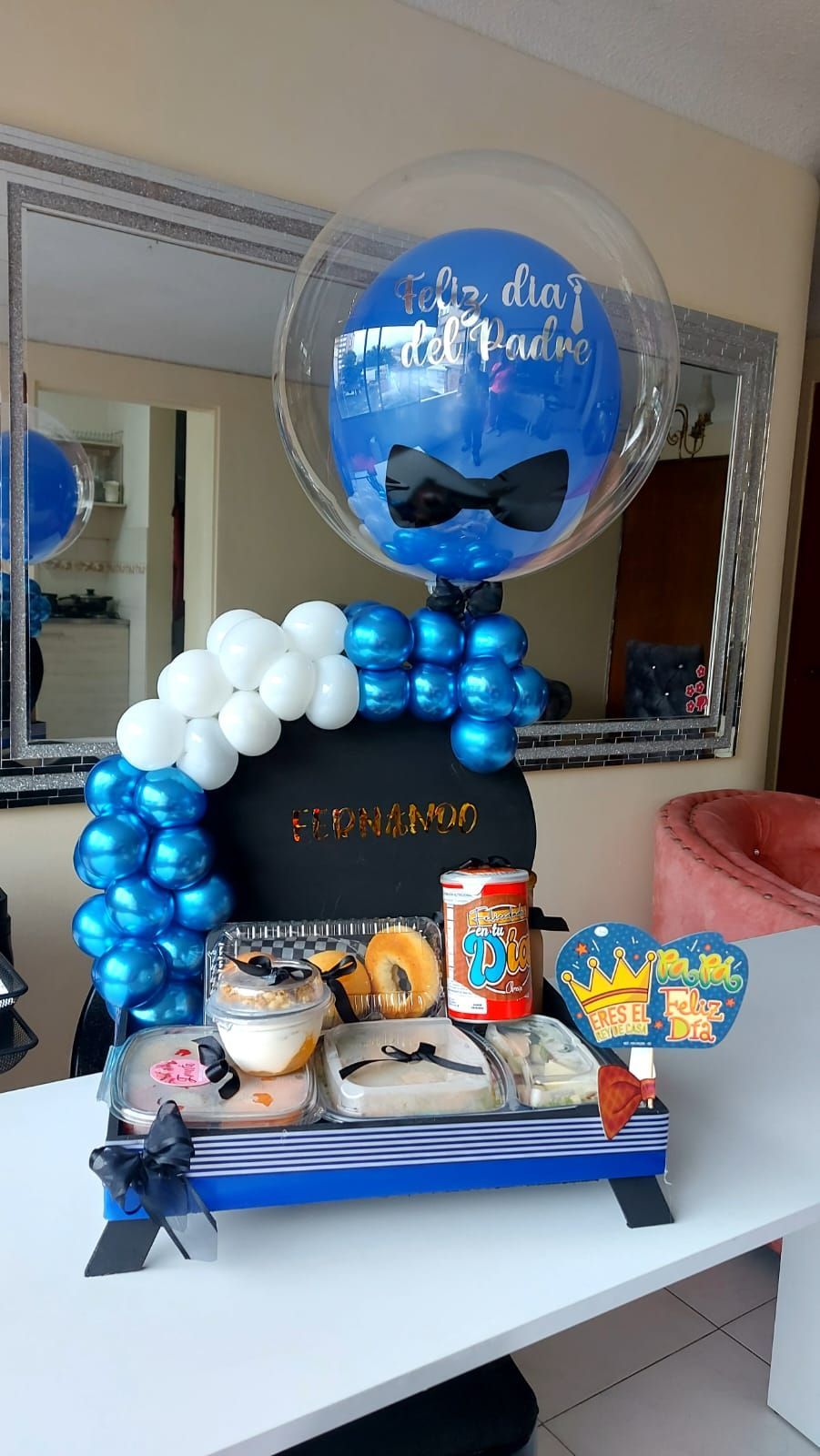 Una canasta de regalo con comida, globos y un globo azul con pajarita y la inscripción