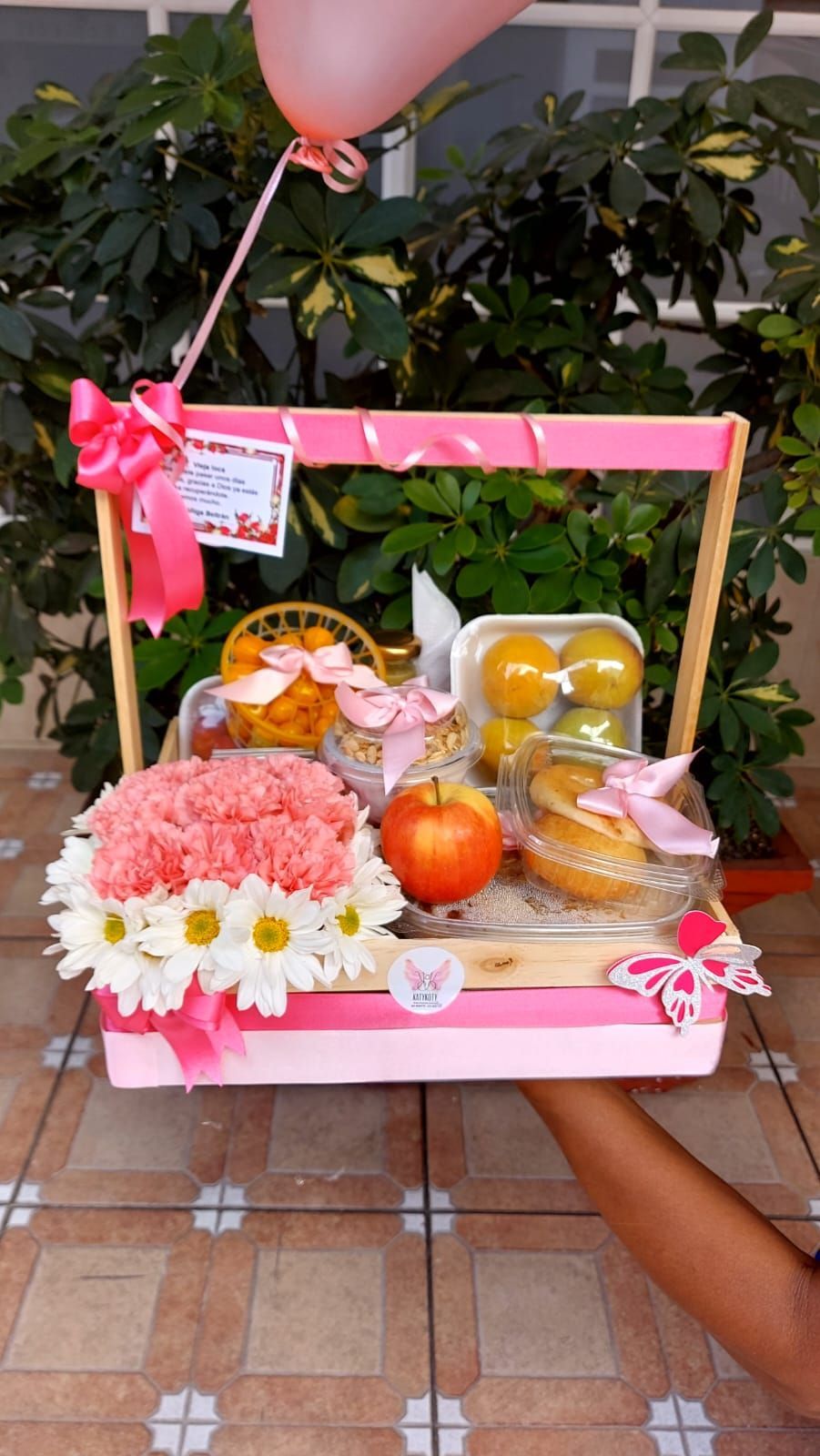 Cesta de regalo rosa con flores, fruta y dulces, colocada al aire libre con un globo rosa.