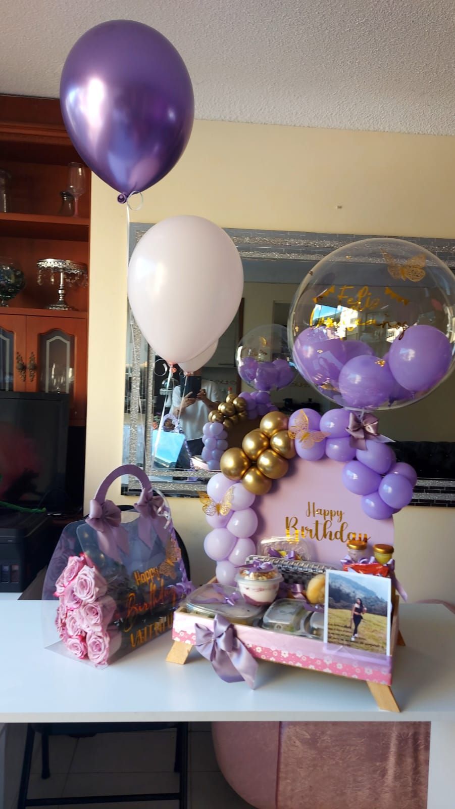 Arreglo de regalo de cumpleaños en tonos morado y dorado con globos, una caja y dulces.