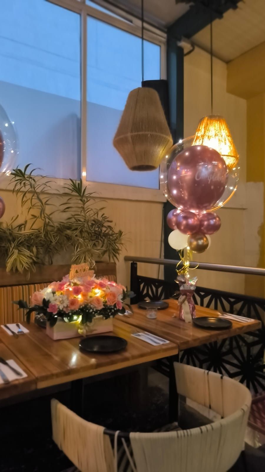 Mesa de restaurante puesta para una celebración. Flores, globos y lámparas colgantes decoran el espacio.