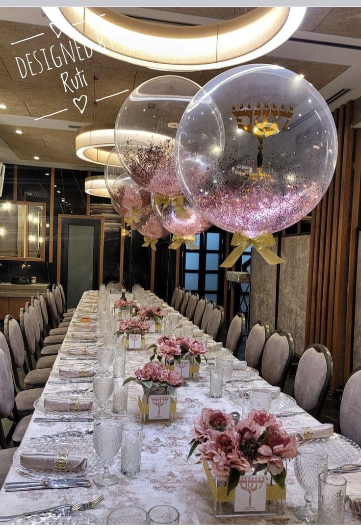Mesa de comedor larga puesta para una celebración, decorada con flores rosas, globos y detalles dorados.