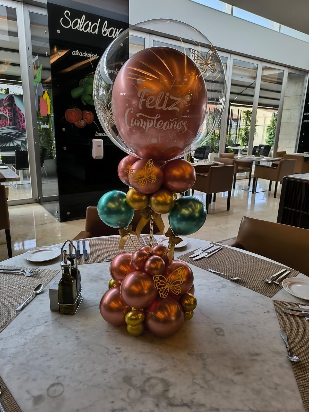 Centro de mesa con globos de cumpleaños en una mesa de restaurante, con globos rosas, dorados y turquesas; texto