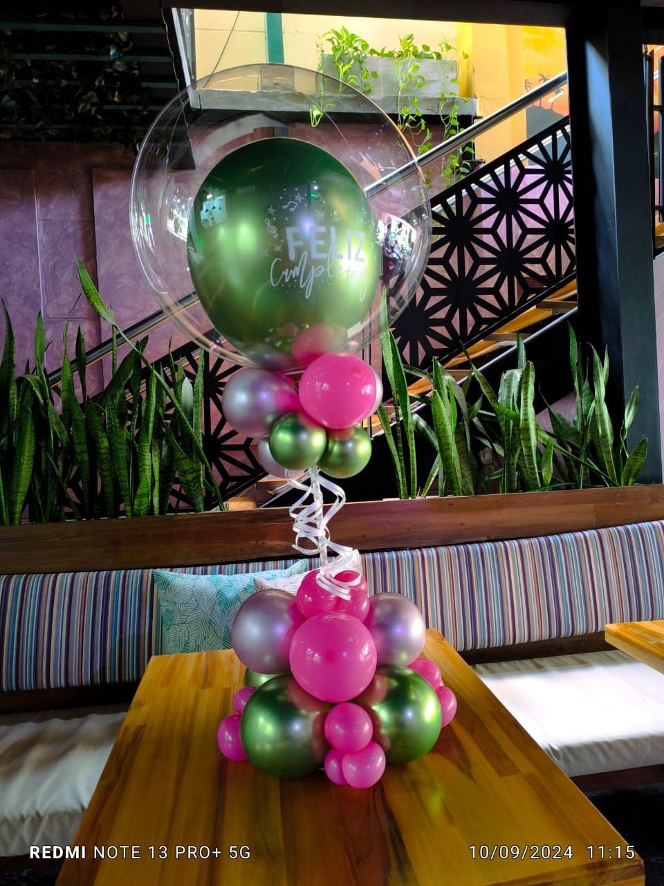 Arreglo floral con globos verdes, rosas y plateados sobre una mesa de madera.