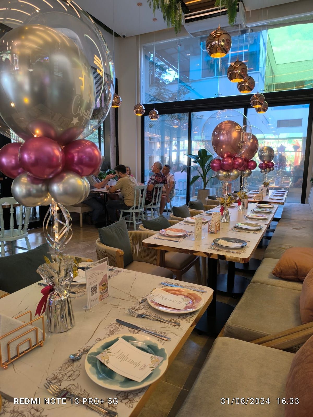 Interior de restaurante con mesas decoradas, globos y gente cenando cerca de un gran ventanal.