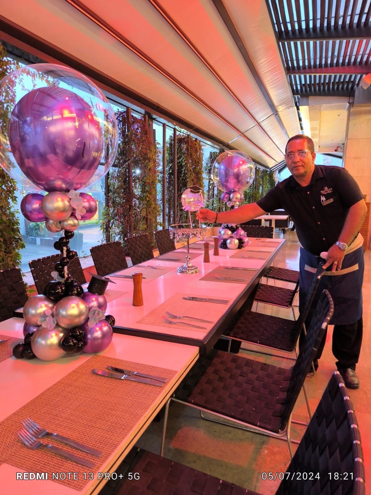 Hombre poniendo la mesa, junto a una mesa larga decorada con globos, en un restaurante.
