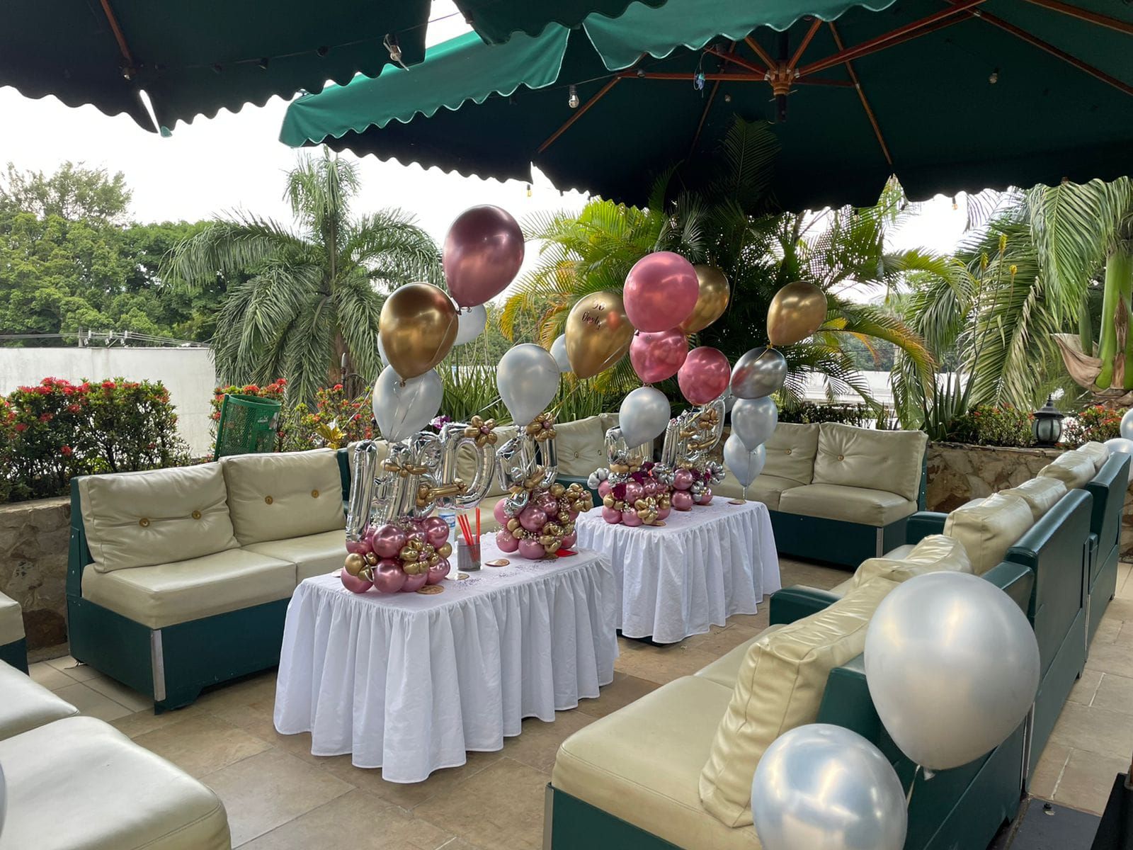 Ambiente de fiesta al aire libre con decoración de globos, mesas blancas y asientos bajo sombrillas verdes.