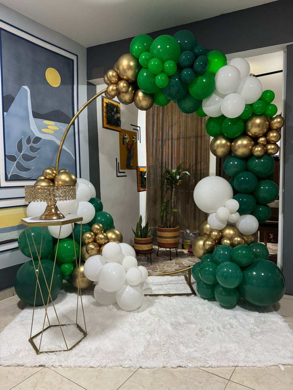 Un arco y arreglos de globos verdes, dorados y blancos decoran una habitación con obras de arte y una alfombra mullida.