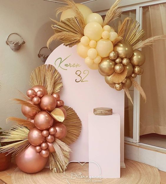 Escenografía de cumpleaños con globos rosas y dorados, hojas de palma secas y un pedestal.
