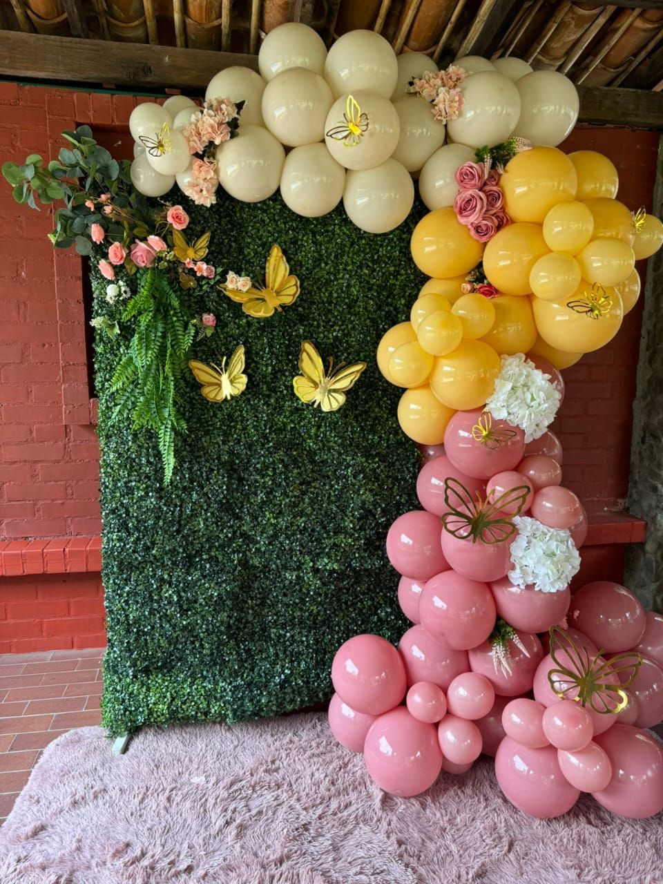 Arco de globos con pared de boj, mariposas y flores en tonos rosa, amarillo y crema.