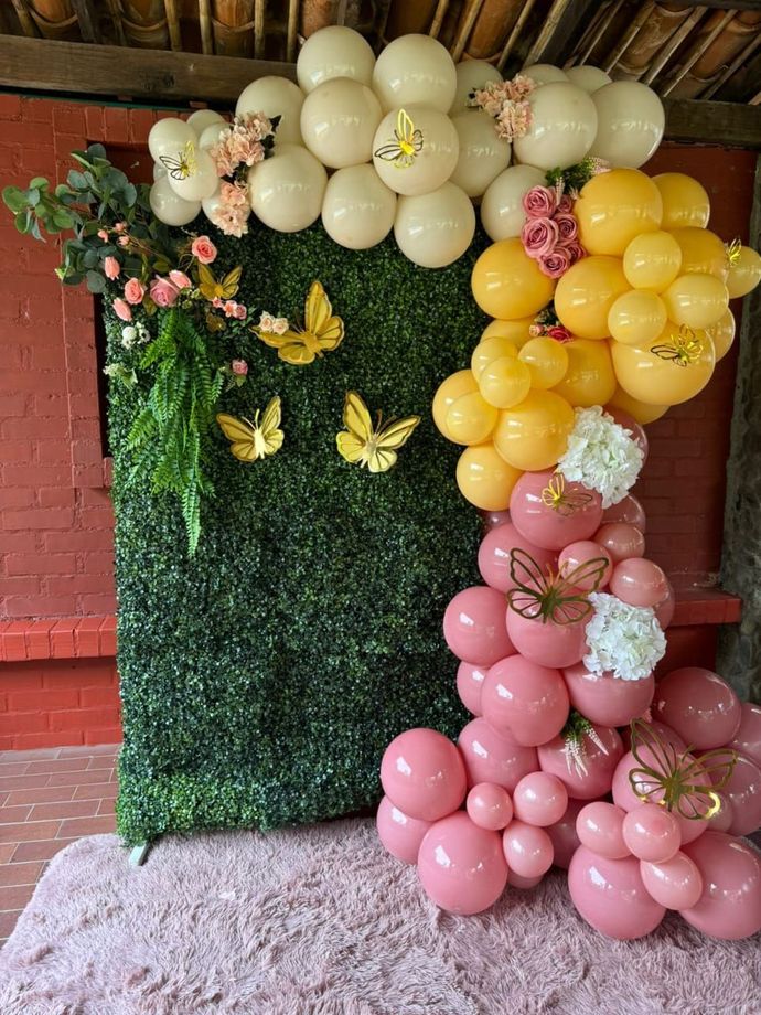 Un muro decorativo de seto verde con detalles de mariposas y un arco de globos rosas, amarillos y crema.