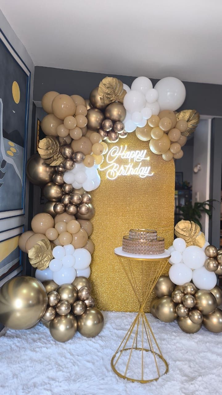 Escenario de cumpleaños con globos dorados y blancos, pared dorada y pastel sobre una mesa pequeña.