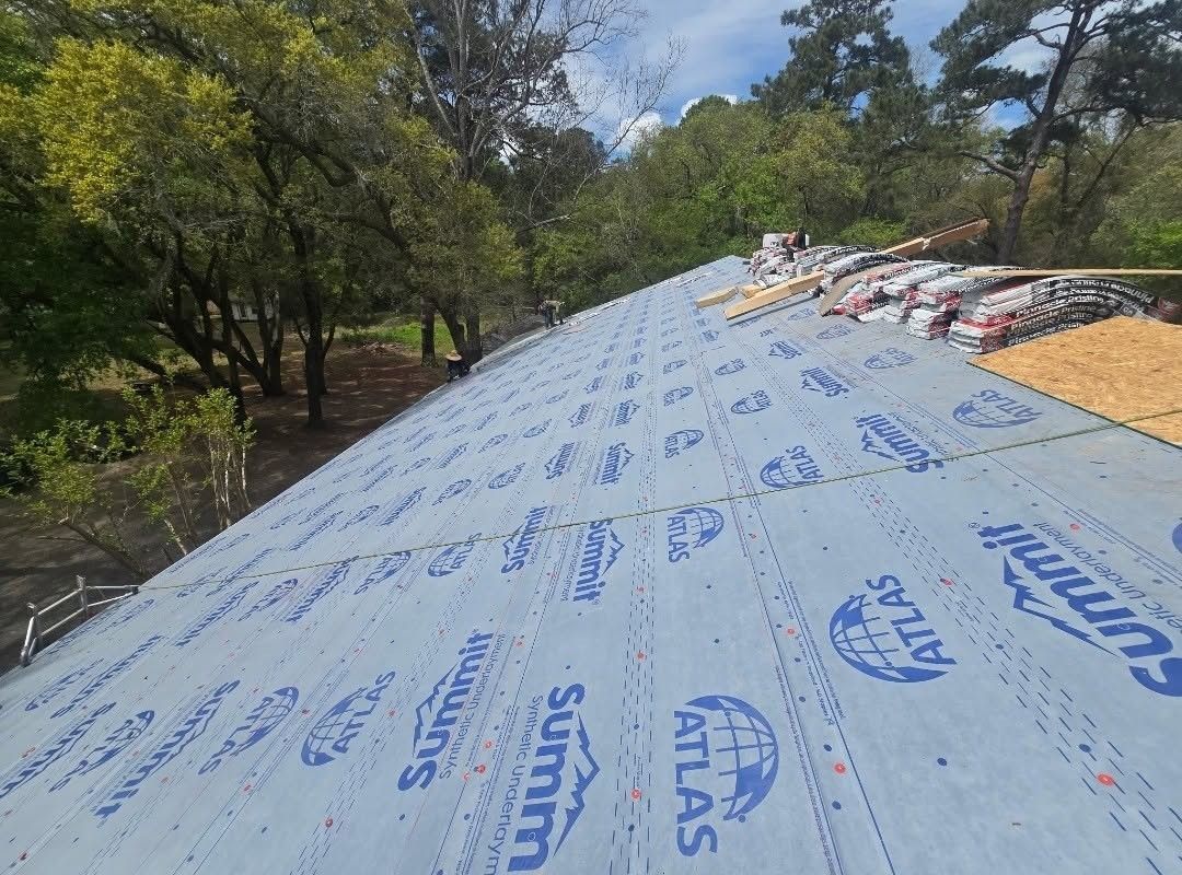 Roof under construction with blue underlayment, lumber stacked at the ridge, and trees surrounding the site