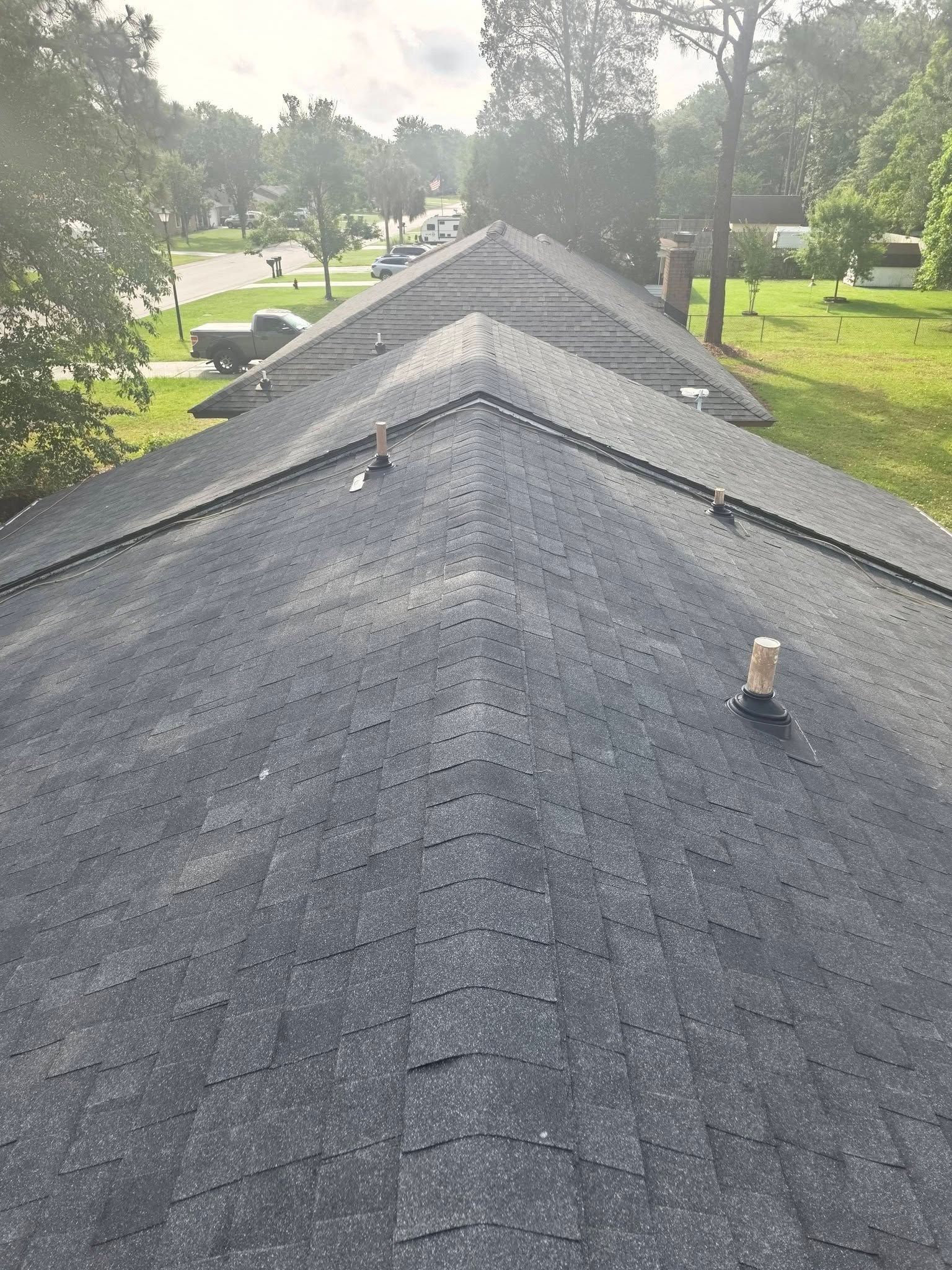 Asphalt shingle roof ridge on a house, viewed from above with trees and neighboring homes in the background.
