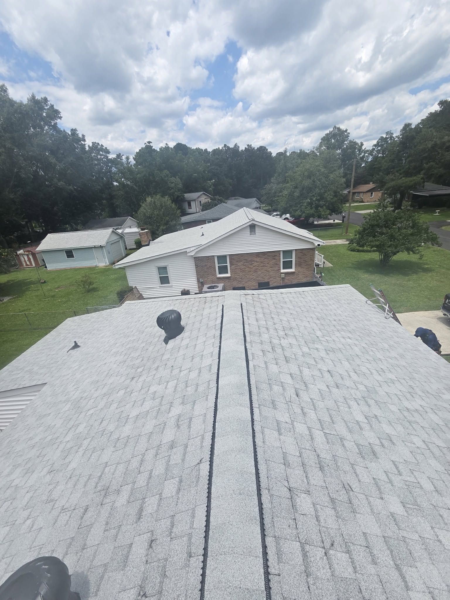 View from a rooftop over a white-shingled house toward a brick home and cloudy suburban neighborhood