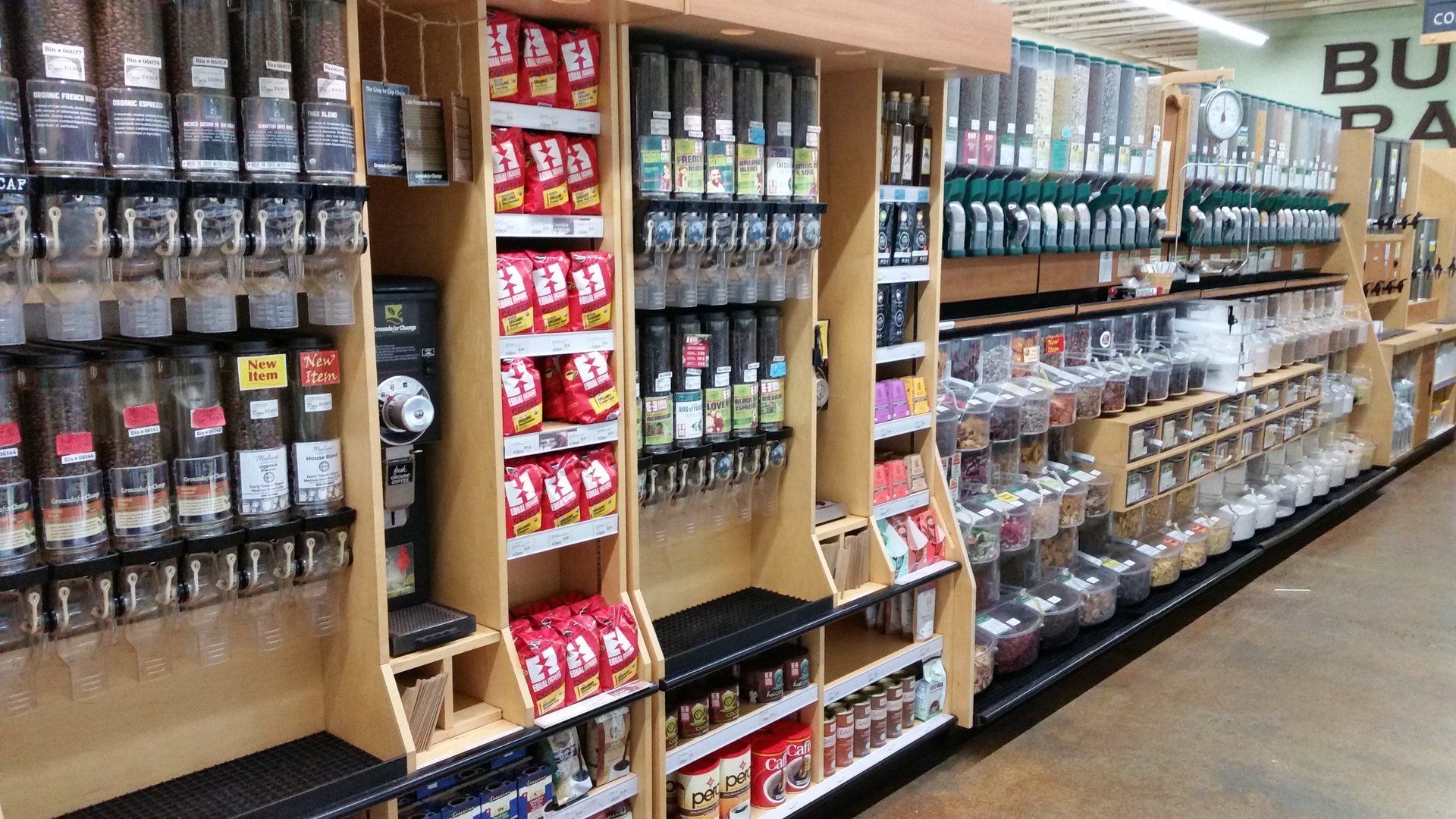 Bulk food dispensers in a store, with clear containers holding grains, beans, and other dry goods. Wooden shelves display packaged products.