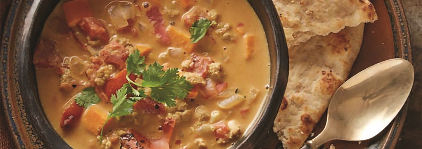 A bowl of soup with bread and a spoon on a rustic plate. The soup is creamy with chunks of meat and vegetables.