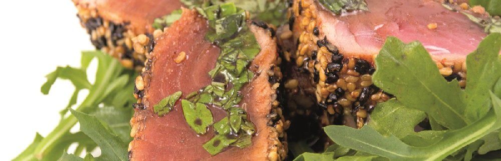 Seared tuna slices coated in sesame seeds, topped with green herbs, on a bed of arugula.
