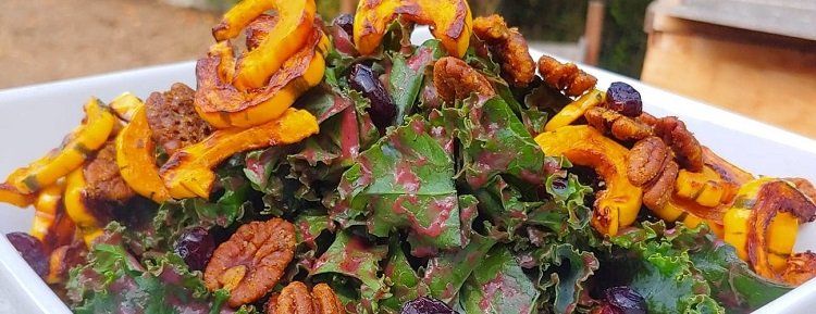 A vibrant salad in a white bowl, featuring colorful greens, orange squash, and pecans. The dish is likely served outdoors, with blurred natural elements in the background.