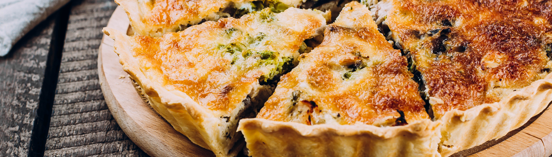 A quiche cut into slices on a wooden serving board. It's golden brown and sits on a wooden table.
