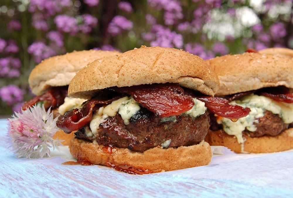 Three gourmet burgers with bacon and blue cheese on a wooden surface, blurred pink flowers in the background.