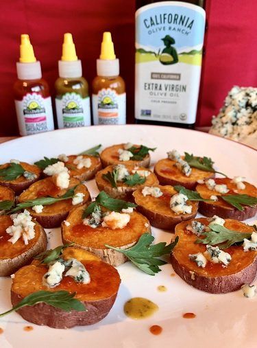 Roasted sweet potato rounds topped with blue cheese, parsley, and hot sauce, with olive oil and hot sauce bottles in the background.