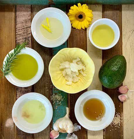 Variety of oils in small bowls, with garnishes and ingredients, on a rustic wooden table. Includes avocado, garlic, and a yellow flower.