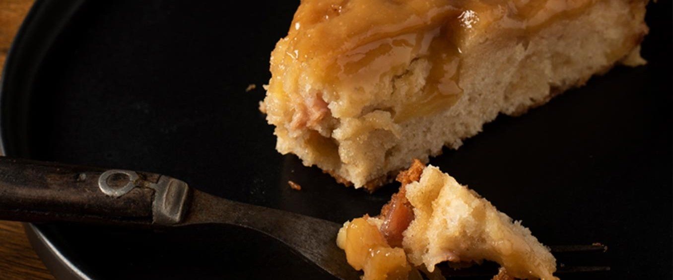 A slice of apple cake on a black plate, a fork holding a piece of the cake. The cake has a glossy caramel topping.