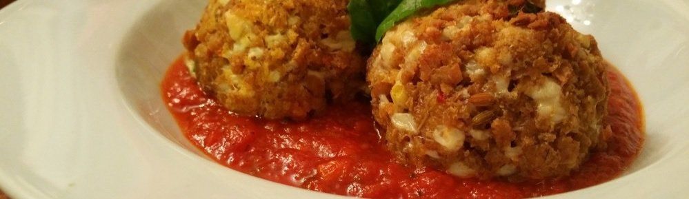 Two breaded balls sit atop a bed of red tomato sauce in a white bowl. Green leaves peek out from between them.