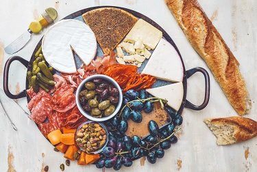 Charcuterie board with various cheeses, meats, olives, grapes, and a baguette on a white wooden surface.
