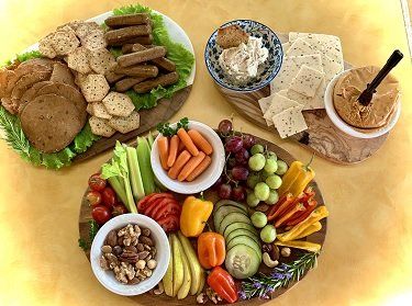 A spread of vegan appetizers on wooden boards: sausages, crackers, dips, vegetables, fruits, and nuts.