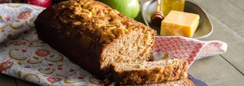 A loaf of apple bread, with a few slices cut off. It sits on a floral cloth next to a green apple and cheese.