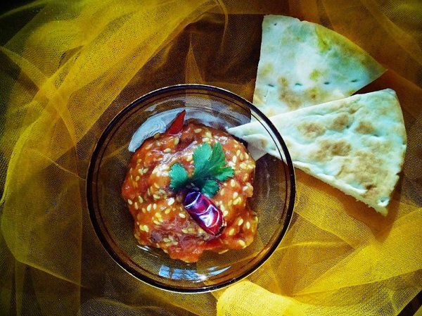Close-up of a small glass bowl filled with a reddish-brown dip, garnished with herbs, alongside flatbread on a yellow fabric.