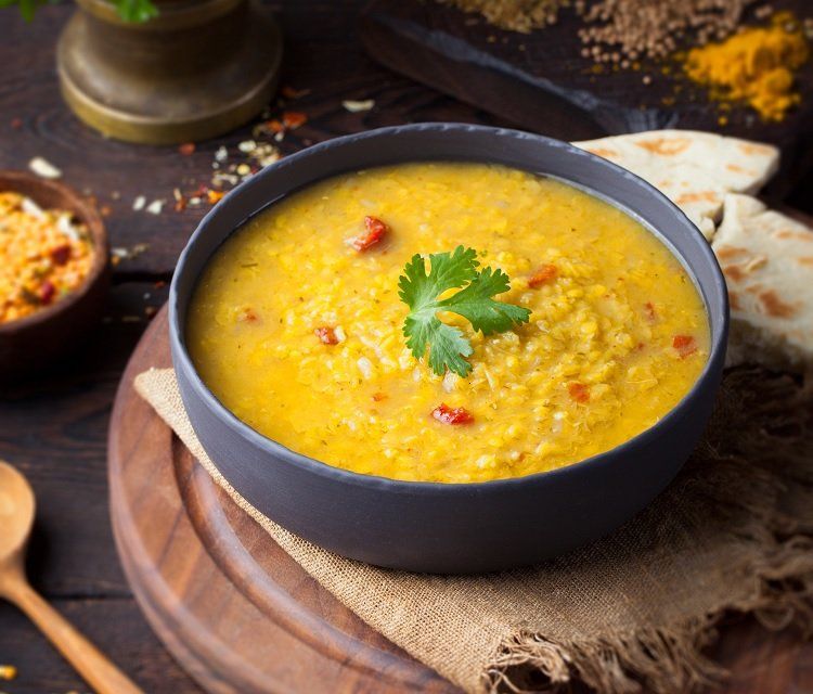 A bowl of yellow lentil soup with a cilantro garnish, served on a wooden board with spices and naan bread.