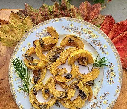 Roasted squash slices with fried sage and rosemary on a floral plate, with fall leaves in the background.