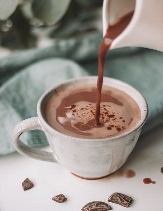 Hot chocolate being poured into a white mug, with chocolate shavings scattered nearby. Soft focus, neutral background.