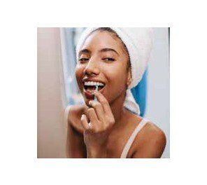 Woman with a towel on her head is flossing her teeth in front of a mirror. She is smiling and the setting is a bathroom.